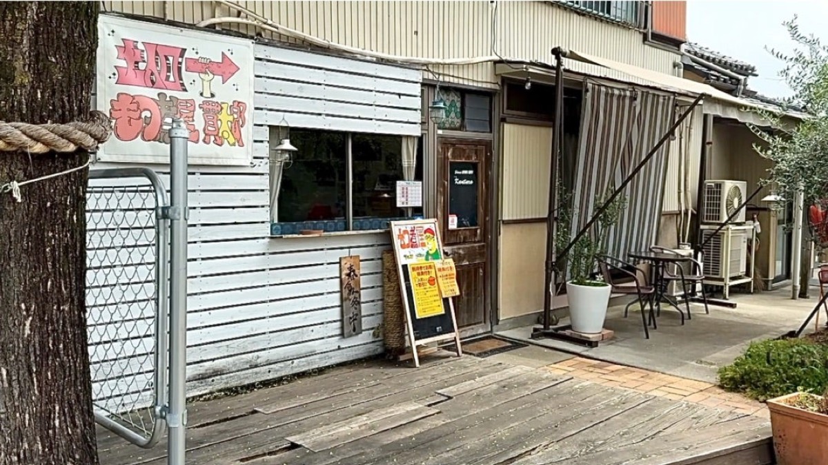 白い下見板張りの外観。手描きの案内看板と木の扉、ウッドデッキの上のテーブルと椅子