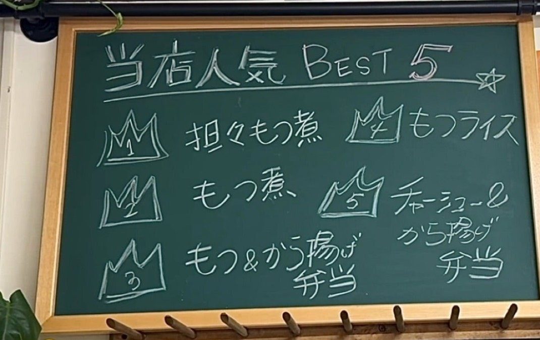 お店の壁の黒板。「当店人気BEST5」として、坦々もつ煮・もつ煮・もつ＆から揚げ弁当・もつライス・チャーシュー＆から揚げ弁当を書いている