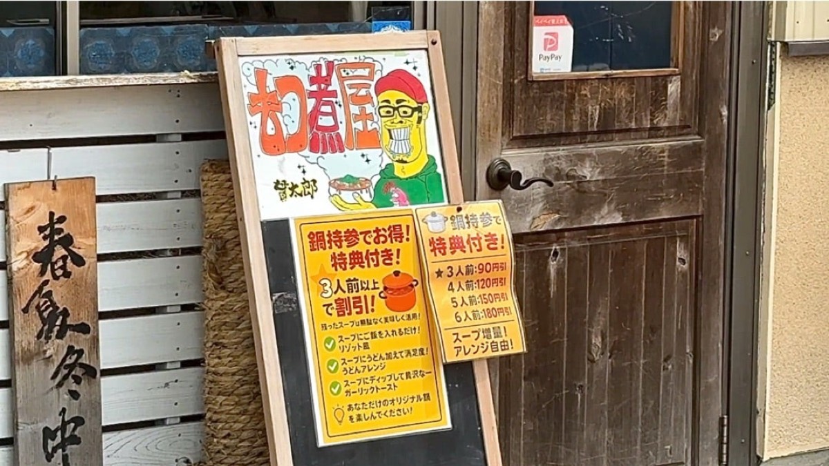 店頭のA型看板。「鍋持参でお得！特典付き！」と書かれた黄色い貼り紙と、木の扉